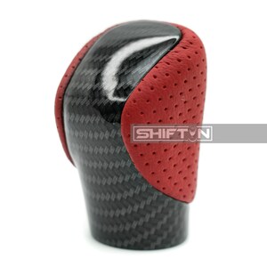 May include: A carbon fiber and red leather shift knob. The shift knob has a glossy black carbon fiber center and red perforated leather sides. The text "SHIFT N" is visible on the side of the shift knob.