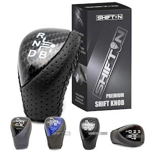 May include: A black and carbon fiber shift knob with the letters R, N, D, and B. The shift knob has a perforated leather grip and is next to a black box with the text "SHIFT N PREMIUM SHIFT KNOB".