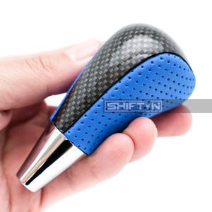May include: A car shift knob with a carbon fiber top and blue perforated leather sides. The shift knob has a silver metal base and the text "SHIFT N" is visible on the side.