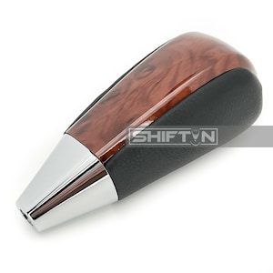 May include: A car gear shift knob with a wood grain top, black leather sides, and a chrome base. The text "SHIFT N" is visible on the side. The knob is designed for automotive use.