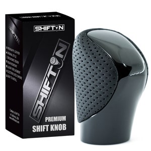 May include: A black gear shift knob featuring a smooth, glossy top section and a textured, perforated grey side panel. The product is displayed next to its black retail box, which is labelled with the brand name SHIFTIN and the text PREMIUM SHIFT KNOB.