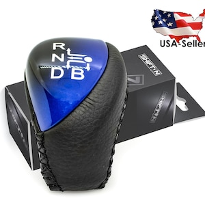 May include: A black and blue car gear shift knob with white lettering indicating the gear positions. The knob is partially wrapped in black leather and sits in front of a black box with the text "SHIFT.VN".