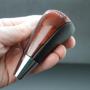 May include: A car gear shift knob with a polished wood grain top and a black leather grip. The base is a shiny chrome finish. The knob is held in a hand.
