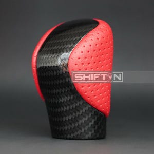 May include: A close-up of a car shift knob. The top is black with a carbon fiber weave pattern, and the sides are red with small perforations. The text "SHIFT N" is visible on the side.