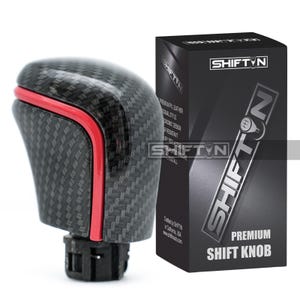 May include: A carbon fiber shift knob with a red accent and a black box. The box has the text "SHIFT N" and "PREMIUM SHIFT KNOB". The shift knob is designed for a vehicle's gear shift.
