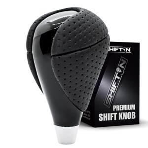 May include: A black gear shift knob featuring a glossy finish and perforated leather-like side panels. The base is silver-toned. Behind the knob stands a black product box with the text SHIFTIN and PREMIUM SHIFT KNOB printed in white.