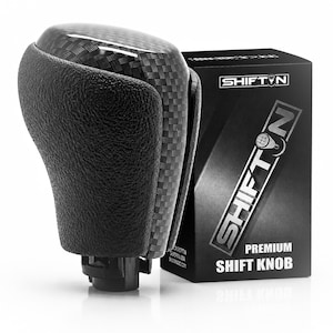 May include: A black gear shift knob featuring a textured leather-like side grip and a glossy carbon fiber top pattern. Next to it stands a black rectangular product box with the white text SHIFTON and the words PREMIUM SHIFT KNOB printed on it.
