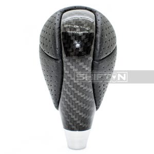 May include: A black and carbon fiber shift knob with a perforated leather grip. The shift knob has a glossy top and a silver base. The text "SHIFT" is visible on the side.