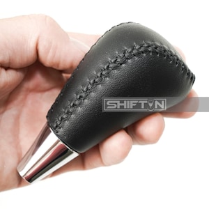 May include: Black leather shift knob with visible stitching and a chrome base. The shift knob is held in a hand and features the text "SHIFT N".