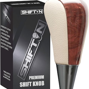 May include: A premium shift knob with a two-tone design. The top section features a combination of wood grain and cream-colored leather, while the base is metallic. The product is packaged in a black box with the brand name "SHIFT N" and product details.