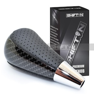 May include: A black and silver shift knob with a carbon fiber design and perforated leather top. The knob has a chrome base and is next to a black box with the text "SHIFTN".