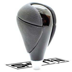 May include: A black and silver car gear shift knob. The top is a glossy black, with a textured black leather grip. The base is a matte black, and the bottom is white. The logo "SHARON" is visible at the base.