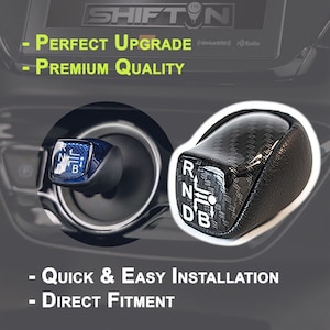 May include: Black carbon fiber gear shift knob with white lettering for a car. The lettering reads "R N D B".