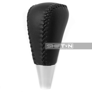 May include: A black leather shift knob with visible stitching. The top portion is rounded and the bottom is a shiny silver color. The text "SHIFT N" is visible on the side.