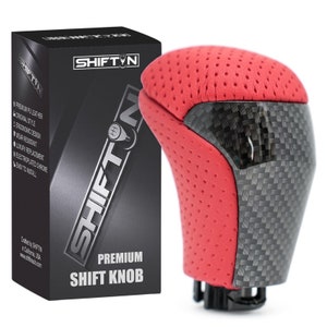 May include: A red and carbon fiber premium shift knob with the text "SHIFT N" and "PREMIUM SHIFT KNOB". The shift knob has a perforated red leather top and a carbon fiber side panel. The product box is black with silver text.