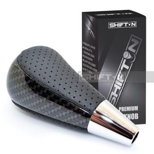 May include: A black and silver shift knob with a carbon fiber pattern and perforated leather top. The knob has a chrome base and is next to a black box with the text "SHIFT N" and "PREMIUM KNOB".