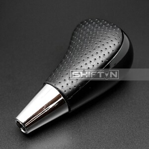 May include: A black and silver car gear shift knob. The top portion is black with a perforated texture. The bottom is silver. The word "SHIFT N" is printed on the side of the knob.