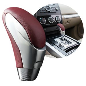 May include: A car gear shift knob featuring a textured deep red leather-like grip accented by smooth silver metallic side panels and a dark metallic base, shown in isolation and installed in a vehicle center console with climate controls.