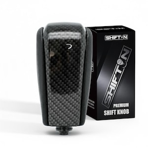 May include: A black and gray shift knob with a carbon fiber weave pattern. The shift knob is next to a black box with the text "SHIFTN" and "PREMIUM SHIFT KNOB" in white.