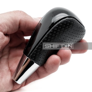 May include: A black and silver car gear shift knob with a perforated leather grip. The top is glossy black, and the base is silver. The text "SHIFT N" is visible on the side.