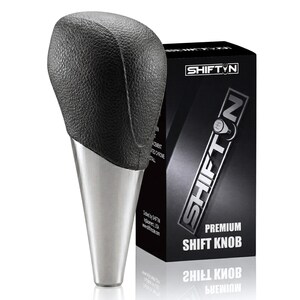 May include: A black textured gear shift knob with a tapered silver metallic base. The knob is positioned next to a black rectangular product box labeled SHIFTN PREMIUM SHIFT KNOB with a graphic of a gear pattern on the letter O.