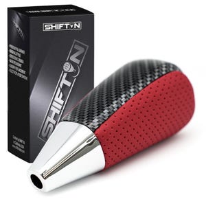 May include: A black and red shift knob with a carbon fiber design and chrome accents. The shift knob is next to a black box with the word "SHIFT" in white.