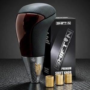 May include: A black and brown shift knob with a leather top and wood grain accents. The shift knob has a silver base with gold detailing and is accompanied by a black box with the text "SHIFT N" and three gold rings.