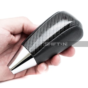 May include: A black and silver car gear shift knob with a carbon fiber top and a silver base. The shift knob has the text "SHIFT N" on the side.