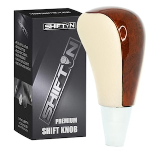 May include: A premium shift knob with a two-tone design. The top half is a rich, dark wood grain, while the lower half is a cream color. The base is white. The box next to it has the text "SHIFT N PREMIUM SHIFT KNOB".