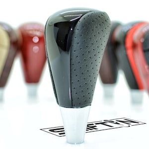 May include: A black and perforated leather shift knob with a glossy black top and a silver base. Other shift knobs in various colors are slightly out of focus in the background. Automotive accessory.