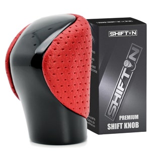 May include: A black and red perforated leather shift knob with a glossy black top section. Next to it stands a black rectangular box with the text SHIFTN and PREMIUM SHIFT KNOB printed in white, featuring a graphic of a gear shift pattern.