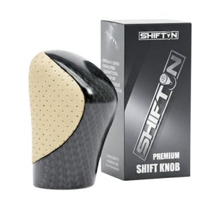 May include: A black and tan shift knob with a carbon fiber pattern and a perforated tan leather grip. The shift knob is next to a black box with the text "SHIFT ON" and "PREMIUM SHIFT KNOB".