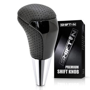 May include: A gear shift knob featuring a perforated dark gray top section, a smooth black center panel, and a polished silver metallic base. Next to it stands a black rectangular box labeled SHIFTON PREMIUM SHIFT KNOB with white text.