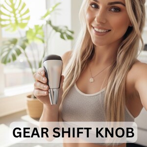 May include: A gear shift knob with a black top, silver body, and chrome base. The text "GEAR SHIFT KNOB" is displayed in a white box at the bottom of the image. The knob is held up by a person.