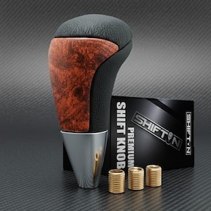May include: A premium shift knob with a black leather top and wood grain accent. The knob has a chrome base and is accompanied by a black box with the text "SHIFT N PREMIUM SHIFT KNOB". Three gold-colored bushings are also included.