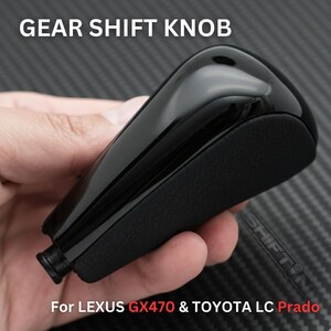 May include: A glossy black gear shift knob with a textured leather-like grip. The text "GEAR SHIFT KNOB" is at the top, and "For LEXUS GX470 & TOYOTA LC Prado" is at the bottom. The knob is held in a hand.