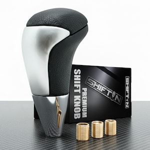 May include: A black and silver shift knob with a leather top and chrome base. The product is accompanied by a black box with the text "SHIFT KNOB PREMIUM" and three gold-colored bushings.