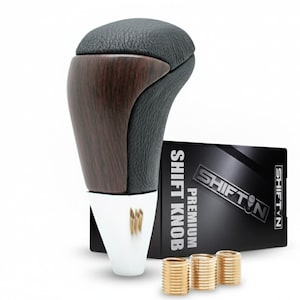 May include: A car shift knob with a dark wood grain finish and black leather top. The knob has a white base and is accompanied by a black box with the text "SHIFT KNOB PREMIUM" and three gold-colored metal rings.