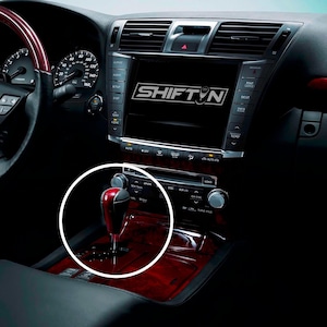 May include: Interior view of a car dashboard featuring a red and black gear shift knob. The dashboard has a wood grain finish and a large screen displaying the word "SHIFTN". The car's interior is dark with various buttons and controls.