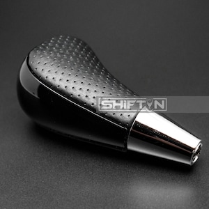 May include: Black and silver car shift knob. The top portion is black with a perforated texture, while the bottom is a shiny silver. The word "SHIFT" is visible on the side.