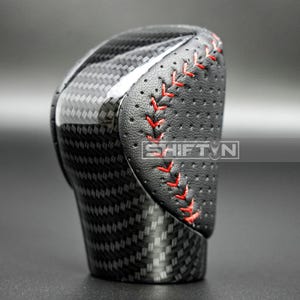 May include: A black and carbon fiber shift knob with red stitching. The shift knob has a perforated leather grip area and the text "SHIFT'N" is visible.