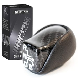 May include: A black and carbon fiber shift knob with the text "SHIFT N" and gear shift indicators. The shift knob is next to a black box with the "SHIFT N" logo and product details.