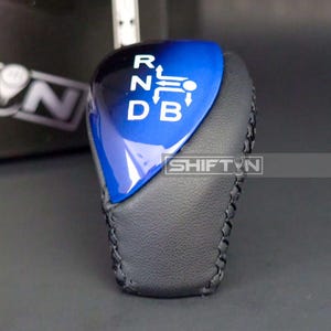 May include: A car gear shift knob with a blue and black top and a black leather base. The top has white lettering indicating the gear positions: R, N, D, B. The brand name "SHIFT'N" is visible.