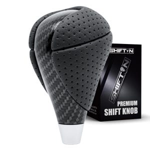 May include: A black gear shift knob featuring a carbon fiber pattern center section flanked by perforated leather-like side panels. The base is a smooth silver-toned metal. The product is displayed next to a black box labeled SHIFTN PREMIUM SHIFT KNOB.