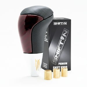 May include: A black and burgundy shift knob with a leather top and a glossy finish. The product is accompanied by a black box with the text "SHIFT N" and three gold-colored metal rings. A white cup with gold designs is also present.