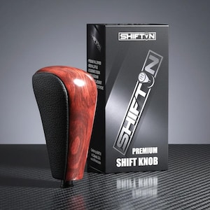 May include: A premium shift knob with a wood grain finish and black leather grip. The product is next to a black box with the text "SHIFT'N" and "PREMIUM SHIFT KNOB". The background is a dark gray.