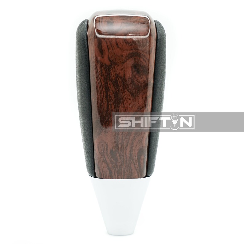 May include: A brown woodgrain shift knob with a black leather boot and a white base. The knob has a glossy finish and a textured woodgrain pattern. The text "SHIFT" is visible on the knob.