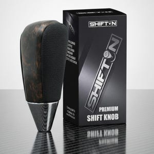 May include: A premium shift knob with a dark wood grain top and black leather grip. The base is chrome. The product is displayed next to a black box with the text "SHIFT'N PREMIUM SHIFT KNOB".