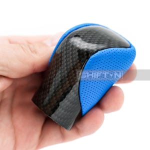 May include: A carbon fiber and blue leather shift knob. The top of the knob is carbon fiber, and the sides are blue leather with small perforations. The text "SHIFTNY" is visible on the side.