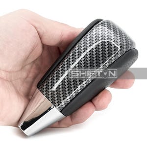 May include: A carbon fiber shift knob with a chrome base. The shift knob has a black leather grip and the text "SHIFT N" is visible on the front.